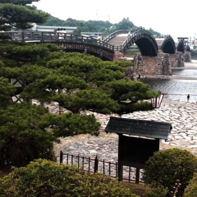 A traditional Japanese wooden arch bridge, Kintai Bridge, spans a river with stone supports.