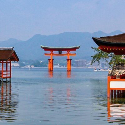 A large red torii gate stands in the water, framed by traditional wooden Japanese buildings with curved roofs, under a blue sky with distant mountains in the background.