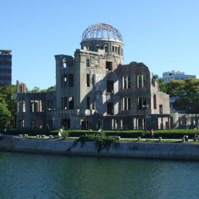 The photo shows the ruins of the Hiroshima Peace Memorial (Genbaku Dome) beside a river under a clear blue sky, surrounded by trees and modern buildings.