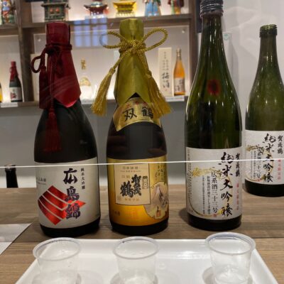 Three bottles of sake with decorative ribbons are displayed behind three empty clear sake cups on a white tray.