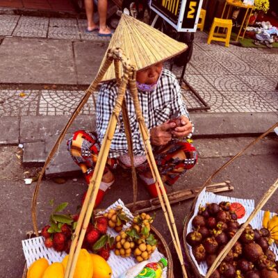 A street vendor wearing a conical hat sits on the sidewalk with baskets of tropical fruits, including mangoes, lychees, strawberries, and mangosteens
