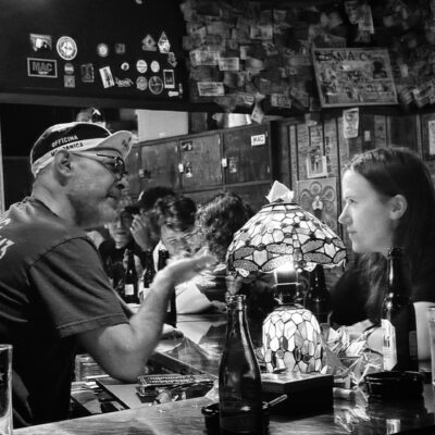Black and white photo of a man and woman talking at a bar, seated with drinks.