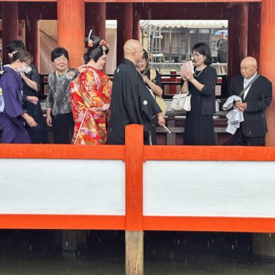 A group of people, including a bride in a red kimono and a groom in black, gather and talk under red pillars during a traditional Japanese wedding ceremony