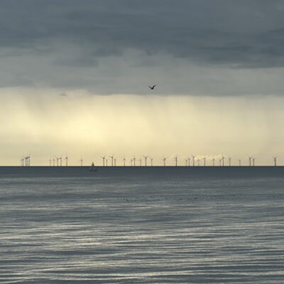 A calm sea under a cloudy sky with distant wind turbines on the horizon.