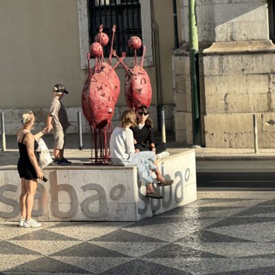 People sit and stand around a stone platform with red abstract sculptures in a sunlit urban setting