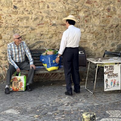 Two men converse by a rustic stone wall; one sits on a bench holding a colorful grocery bag, while the other stands wearing a cowboy hat.
