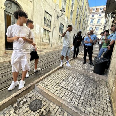 Several young men stand and talk on a narrow cobblestone street with tram tracks, while two police officers and others stand nearby