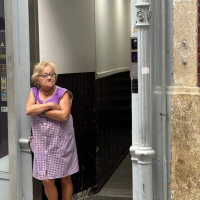 An older woman in a purple checkered dress and red sneakers stands with arms crossed in a doorway, looking to the side