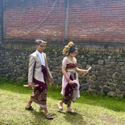Two people in traditional Balinese attire walk side by side outdoors near a stone and brick wall