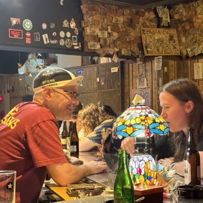Two people sit at a bar covered in bottles and glasses, talking closely beneath a colorful stained glass lamp.