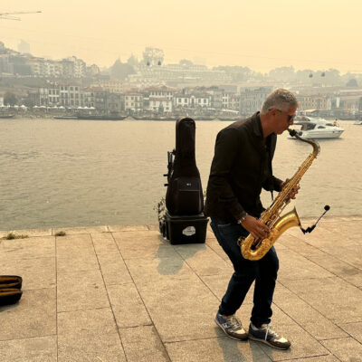 A man plays a saxophone beside a river, with buildings and boats visible in the background