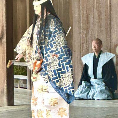 A performer in ornate traditional Japanese attire and mask stands on a wooden stage, holding a fan, while a seated man in blue robes watches
