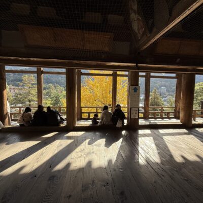 People sit by large wooden windows, enjoying sunlight and views of autumn trees with vibrant yellow leaves