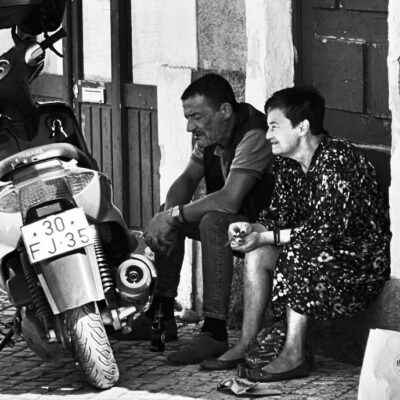 A man and a woman sit on a doorstep next to a parked scooter, deep in conversation with the woman smiling. Sunlight casts strong shadows on the cobblestone street