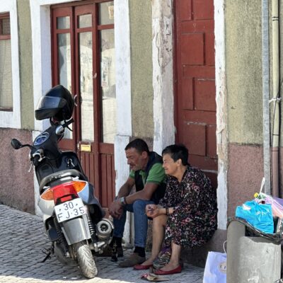 A man and a woman sit together in a doorway next to a parked scooter on a cobblestone street