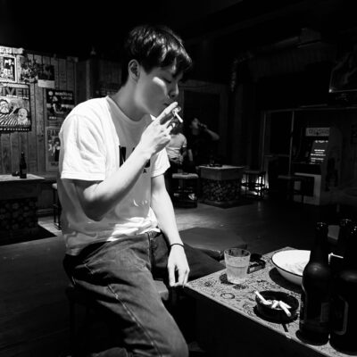 A young man sits at a table in a dimly lit bar, smoking a cigarette. Several bottles, glasses, and an ashtray are on the table
