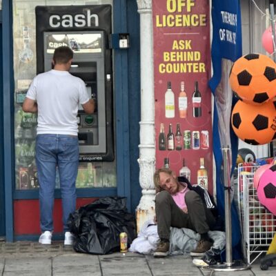 A man uses an ATM outside a shop, while another man sits on the ground nearby surrounded by bags and a can