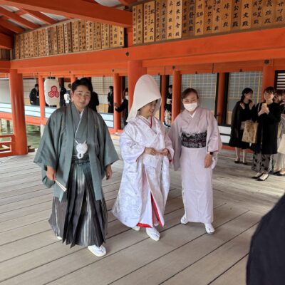 A bride in a traditional white kimono and hood walks with two attendants in kimonos at a Japanese wedding ceremony in a wooden, red and orange pavilion