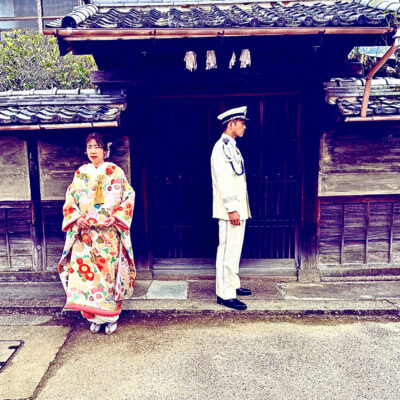 A woman in a colorful kimono stands beside a man in a white military uniform, both facing away from each other in front of a traditional wooden Japanese building